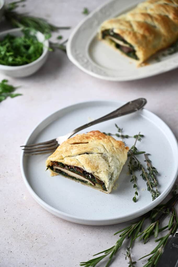 A plate with a slice of mushroom wellington and a serving dish behind with fresh rosemary scattered around.