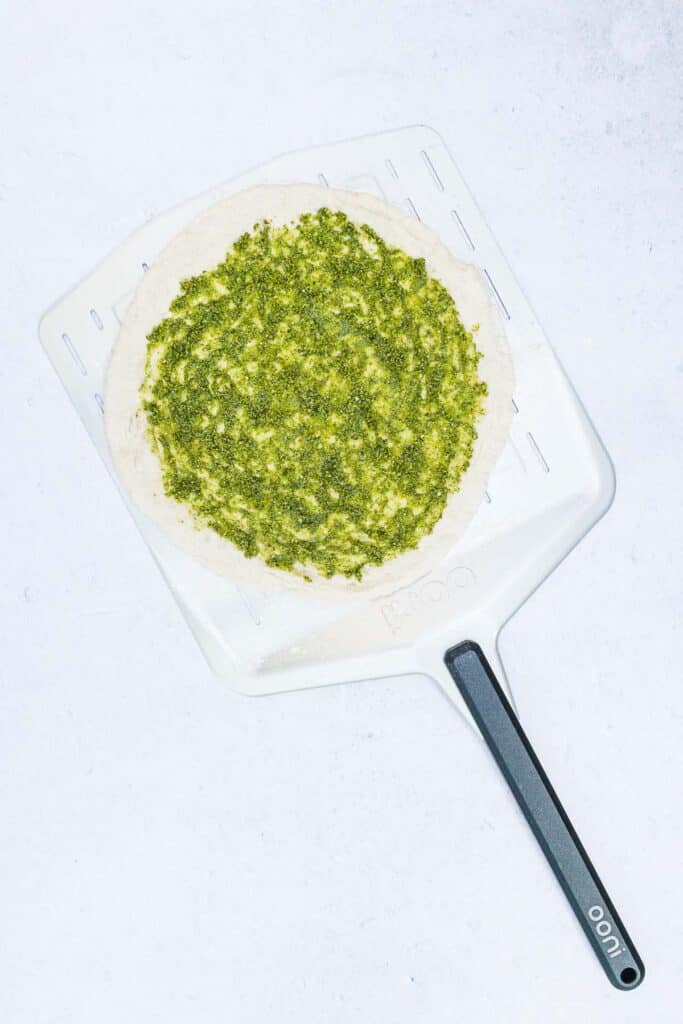 Pizza crust with pesto sauce spread on it resting on a pizza peel.