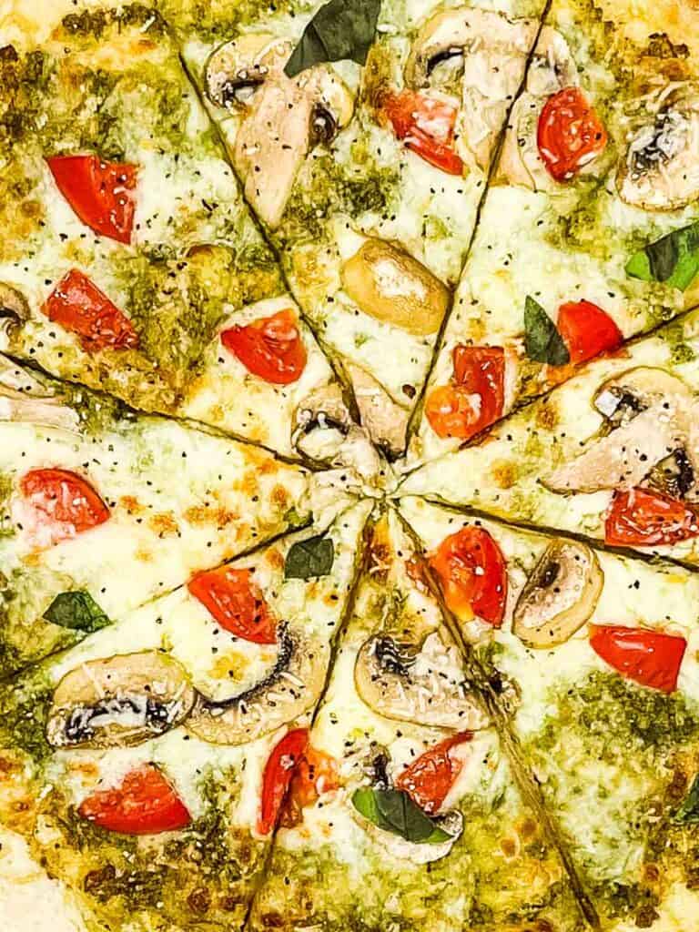 A pizza that is sliced, with pesto, cheese, tomatoes, and mushrooms.