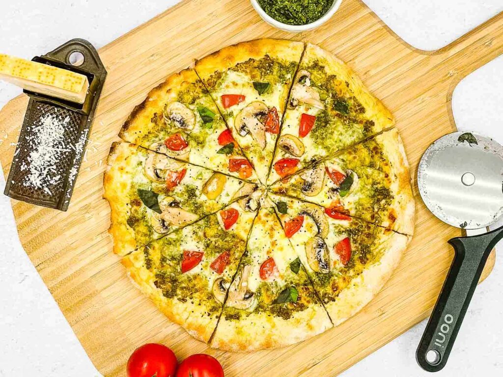 A pesto pizza on a wooden board, pizza cutter, basil pesto, tomatoes, and vegetarian shredded Parmesan cheese in a cheese grater.