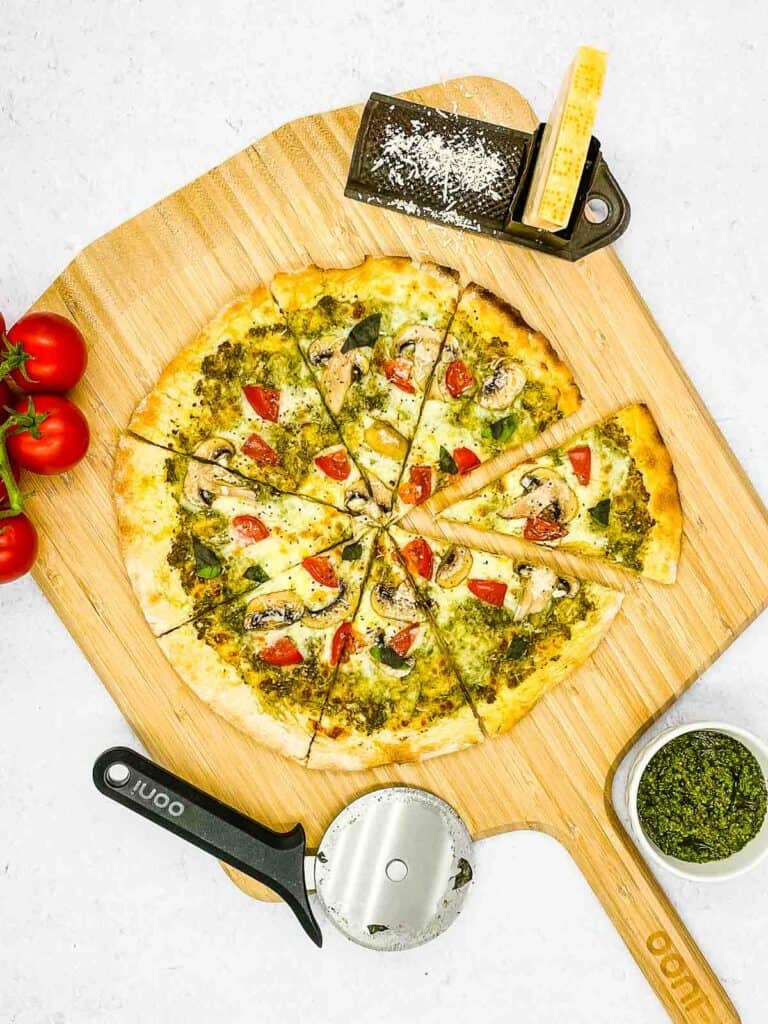 A pizza paddle with a fresh pesto pizza on, a pizza cutter, basil pesto sauce, vegetarian Parmesan in a grater, and tomatoes.