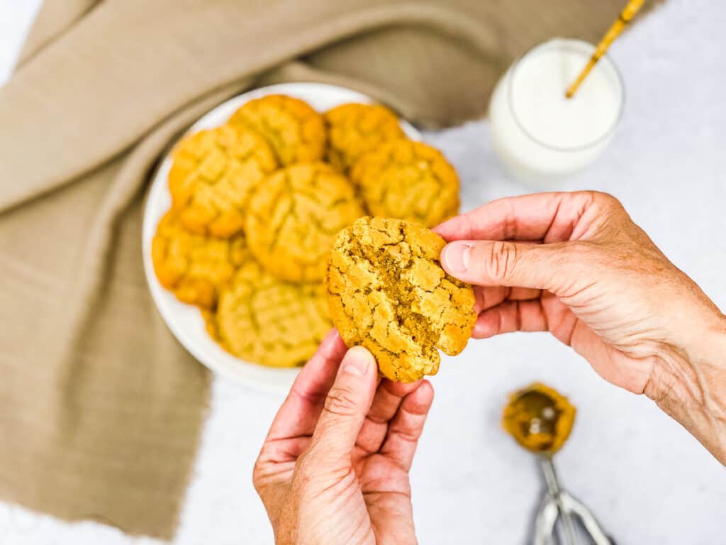 Someone pulling a peanut butter cookie in half, with a plate of them below.