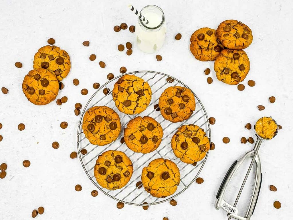 A wire rack with peanut butter chocolate chip cookies on, cookies and chocolate chips scattered around, with milk and a scoop.