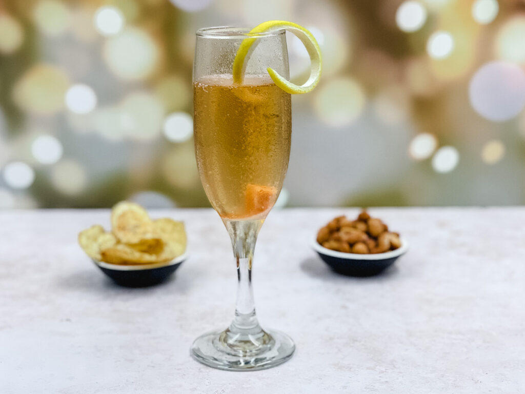 A champagne cocktail, garnished with lemon twist, with nuts and crisps in the background.
