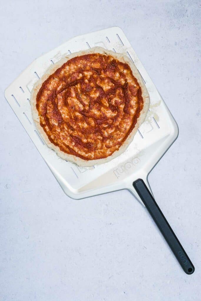 A pizza crust on a pizza peel with marinara sauce on it.