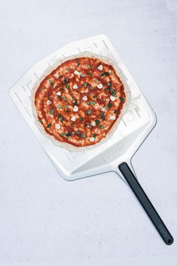A pizza crust on a pizza peel with marinara sauce, fresh thyme, and slices of garlic.