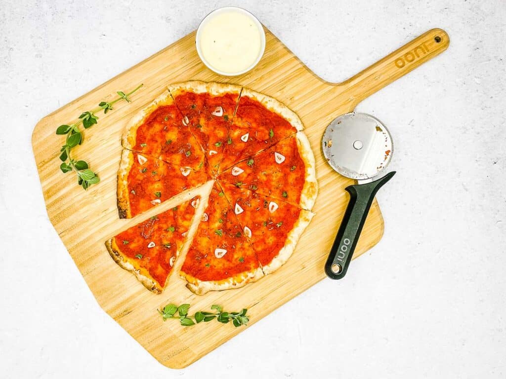 A marinara pizza sliced and on a board, with a pizza cutter, and pizza dipping sauce.