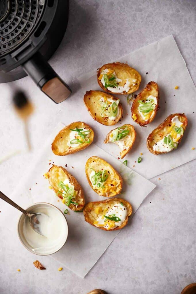 Overhead shot of crispy air fryer potato skins topped with melted cheese, sour cream, and sliced green onions, arranged on parchment paper.