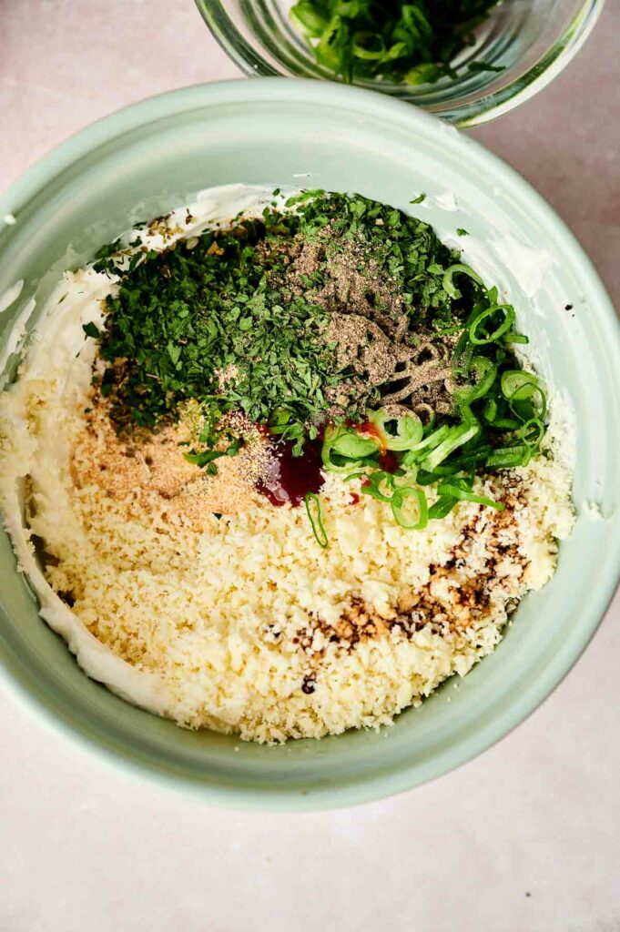 A bowl of cream cheese mixture with herbs, cheese, green onion, and seasonings being combined.