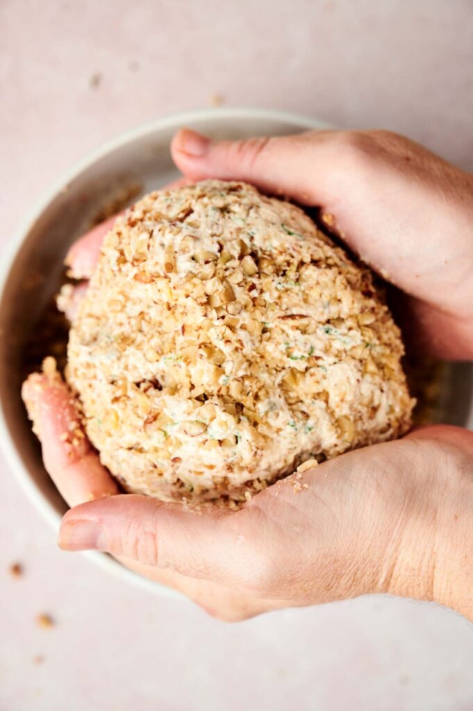 A person holding a ball of mixture in a bowl while preparing a cheese ball recipe.