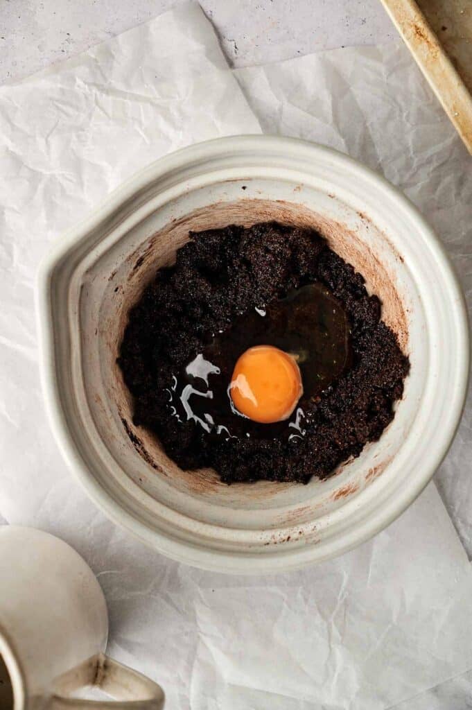 A white mixing bowl contains dark chocolate brownie batter with a raw egg on top, placed on crumpled parchment paper, ready to transform into delectable chocolate crinkle cookies.