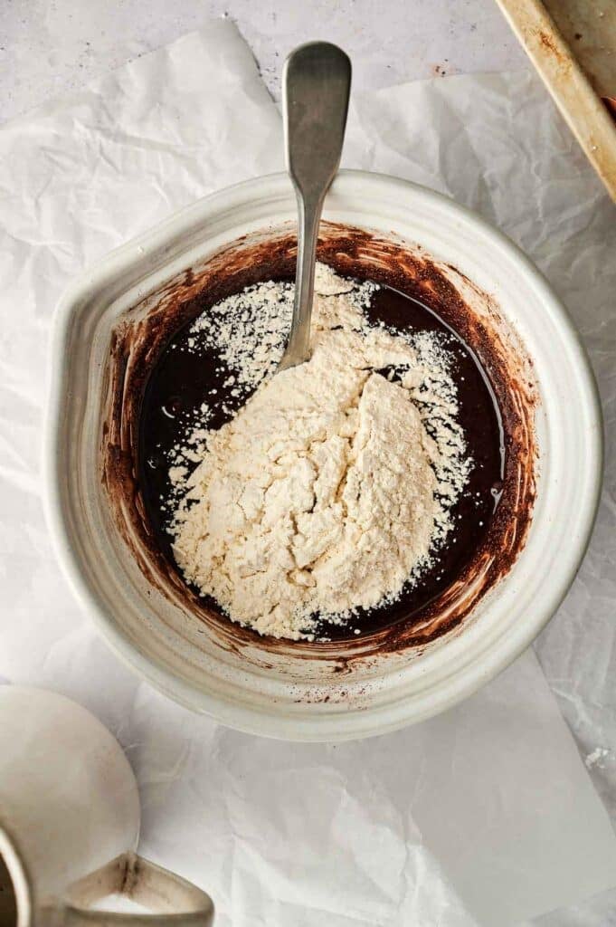 A white mixing bowl with a silver spoon contains a blend of dark brown liquid batter and unmixed flour, hinting at the creation of chocolate crinkle cookies. White parchment paper and light-colored items are visible in the background.