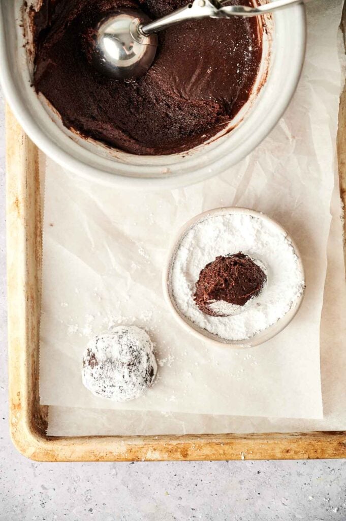 A bowl with chocolate dough and a scoop, a small dish of powdered sugar with a chocolate dough ball inside, and a powdered sugar-coated dough ball on parchment paper—perfect for making delectable chocolate crinkle cookies.