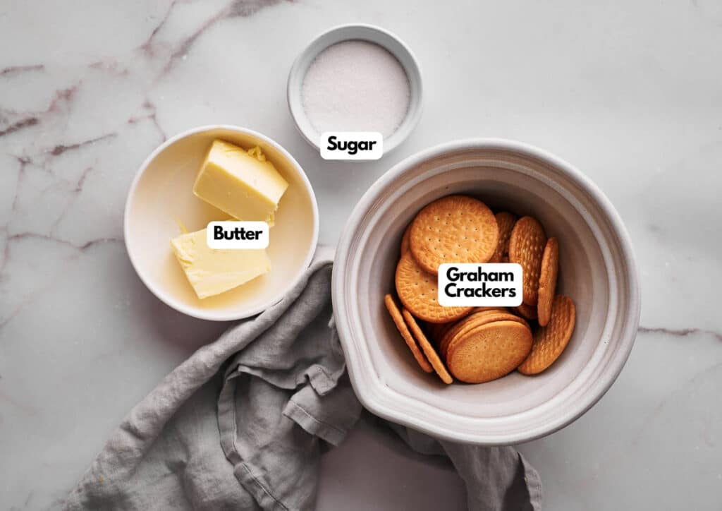 A bowl of graham crackers, a bowl of butter, and a small bowl of sugar are arranged on a marble surface with a gray cloth, perfect for making a classic graham cracker crust.