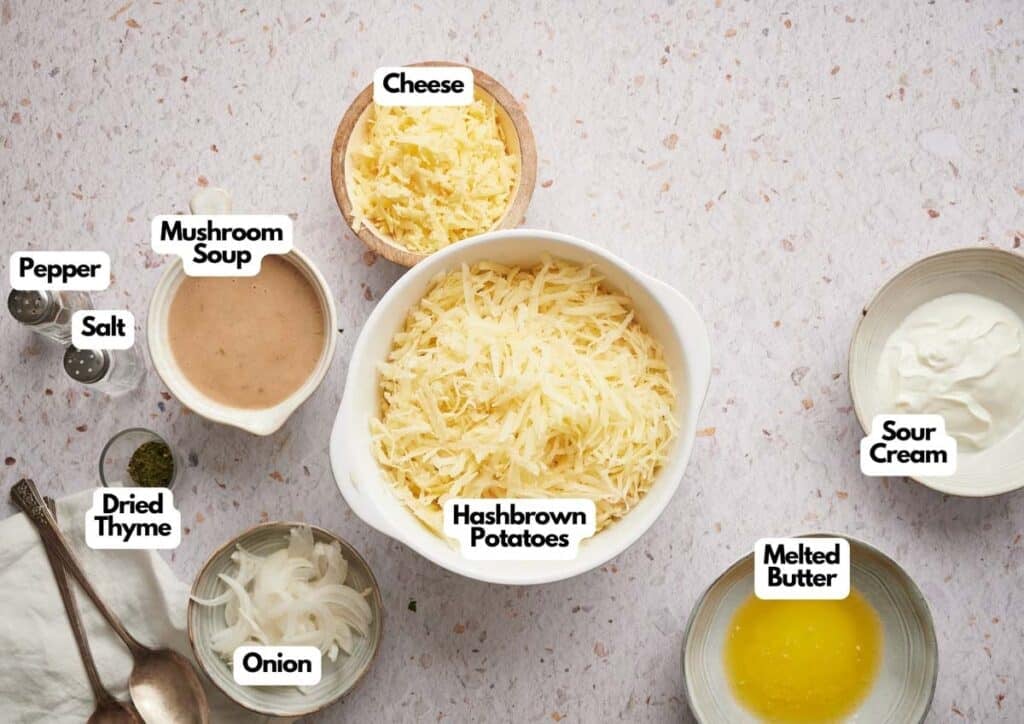 A bowl of shredded hashbrown potatoes surrounded by labeled ingredients for hashbrown casserole: cheese, mushroom soup, sour cream, melted butter, onion, dried thyme, salt, and pepper.