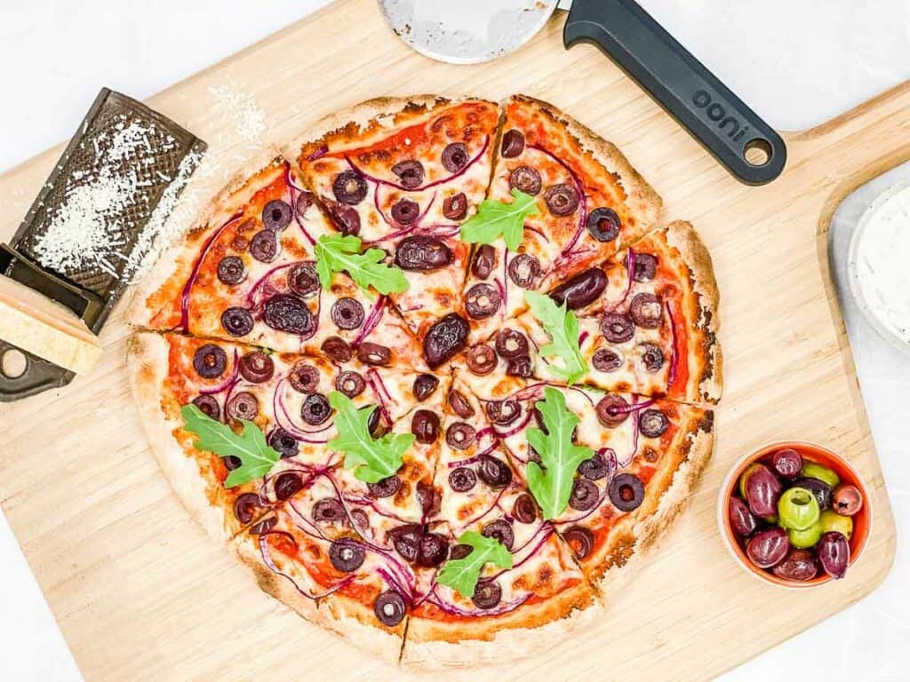 Pizza with olives on wooden peel, with a pizza cutter, olives, and cheese.