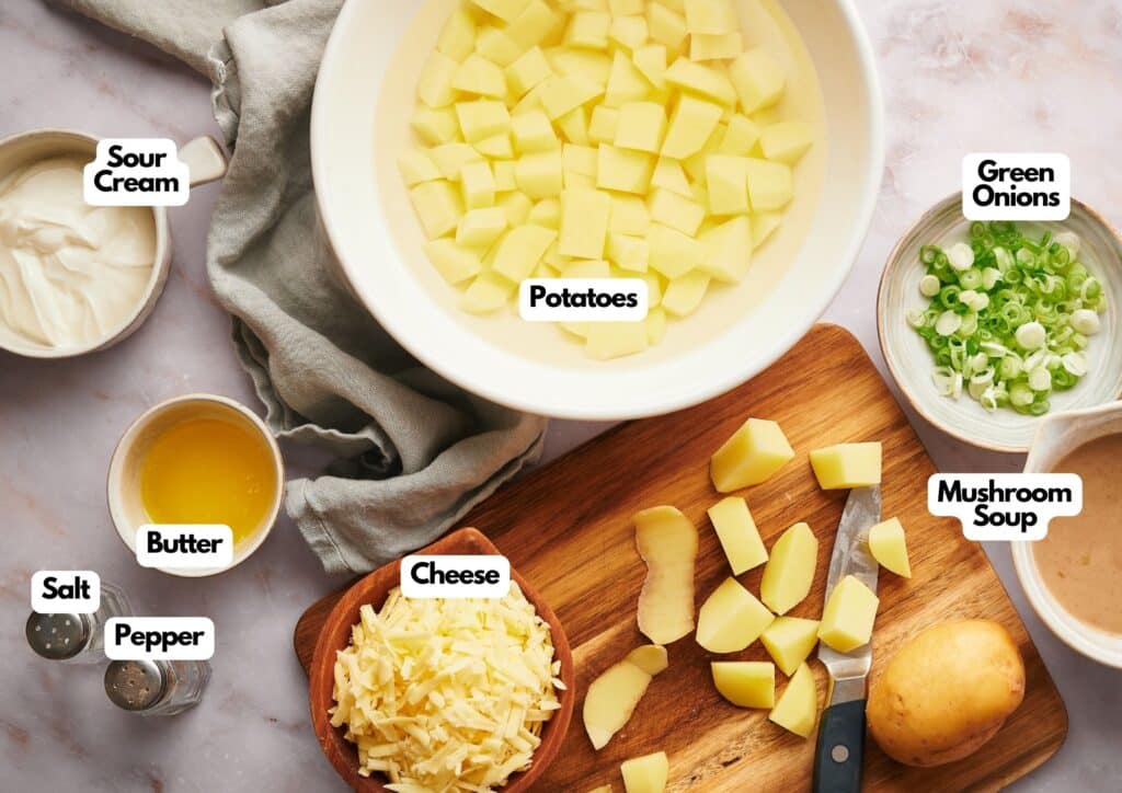 Chopped potatoes in a bowl are ready to be transformed into a delicious potato casserole, surrounded by shredded cheese, sliced green onions, butter, sour cream, mushroom soup, salt, and pepper on a countertop.