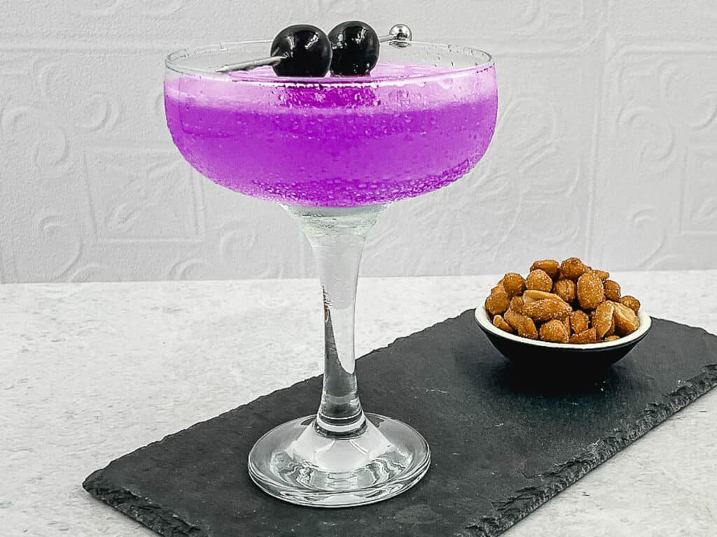 A stunning aviation cocktail on a slate with a bowl of nuts.