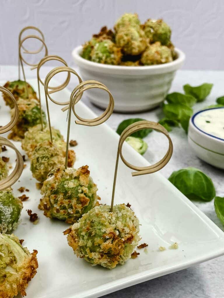 Cheesy Brussels sprouts bites with skewers stuck into them and a bowl of sprouts behind.