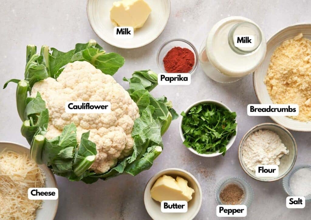 A variety of labeled ingredients on a countertop, perfect for making cauliflower casserole: cauliflower, cheese, butter, milk, paprika, breadcrumbs, flour, pepper, salt, and parsley.