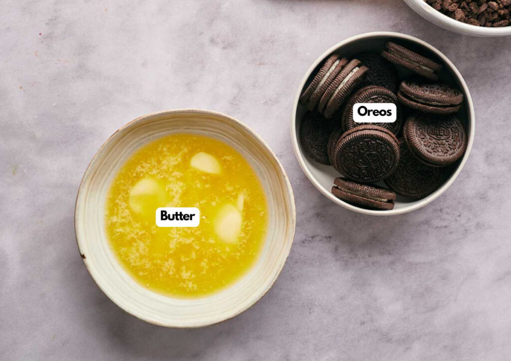 A bowl of melted butter and a bowl of Oreo cookies, essentials for making Mississippi Mud Pie, are placed on a light-colored surface, each labeled accordingly.
