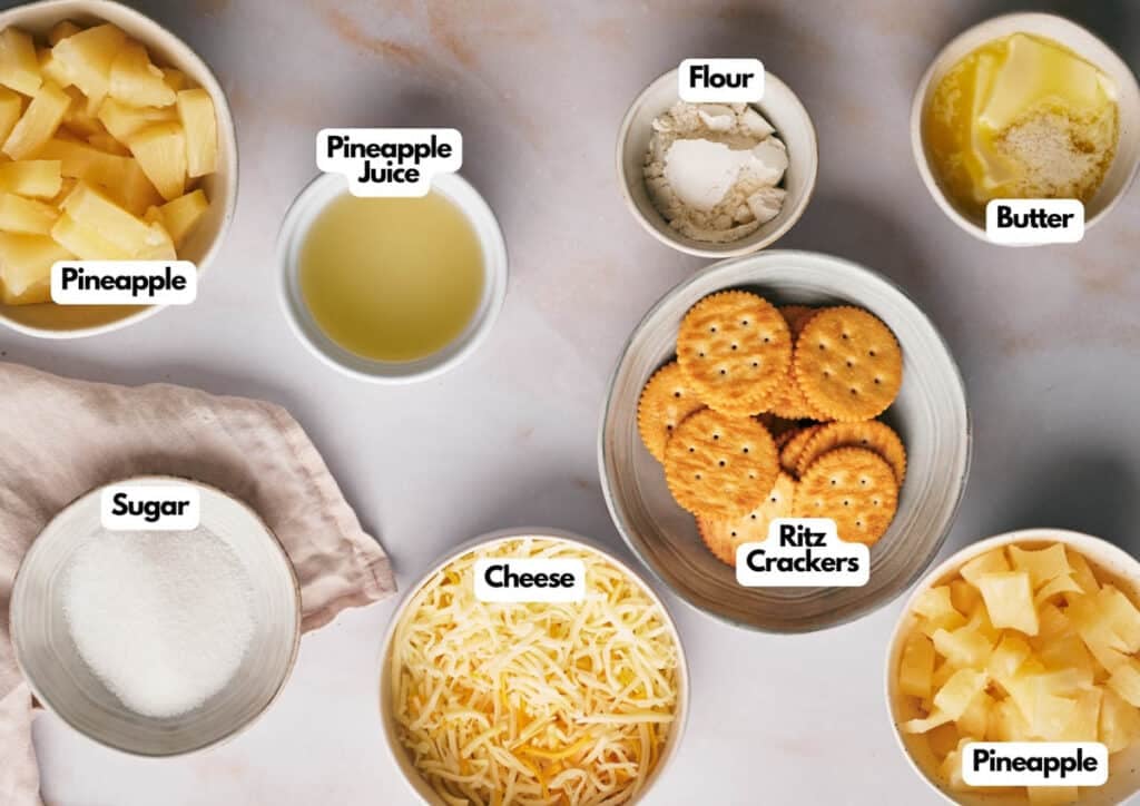 A countertop displays the labeled ingredients for pineapple casserole: bowls of pineapple chunks, pineapple juice, flour, melted butter, sugar, shredded cheese, and Ritz crackers.