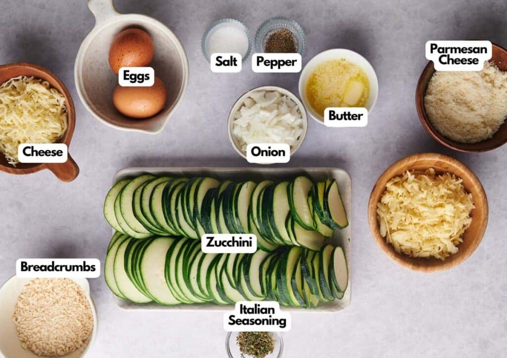 A top-down view of bowls containing eggs, salt, pepper, butter, parmesan cheese, cheese, breadcrumbs, onion, Italian seasoning, and sliced zucchini&mdash;everything you need for a delicious zucchini casserole&mdash;all neatly labeled.