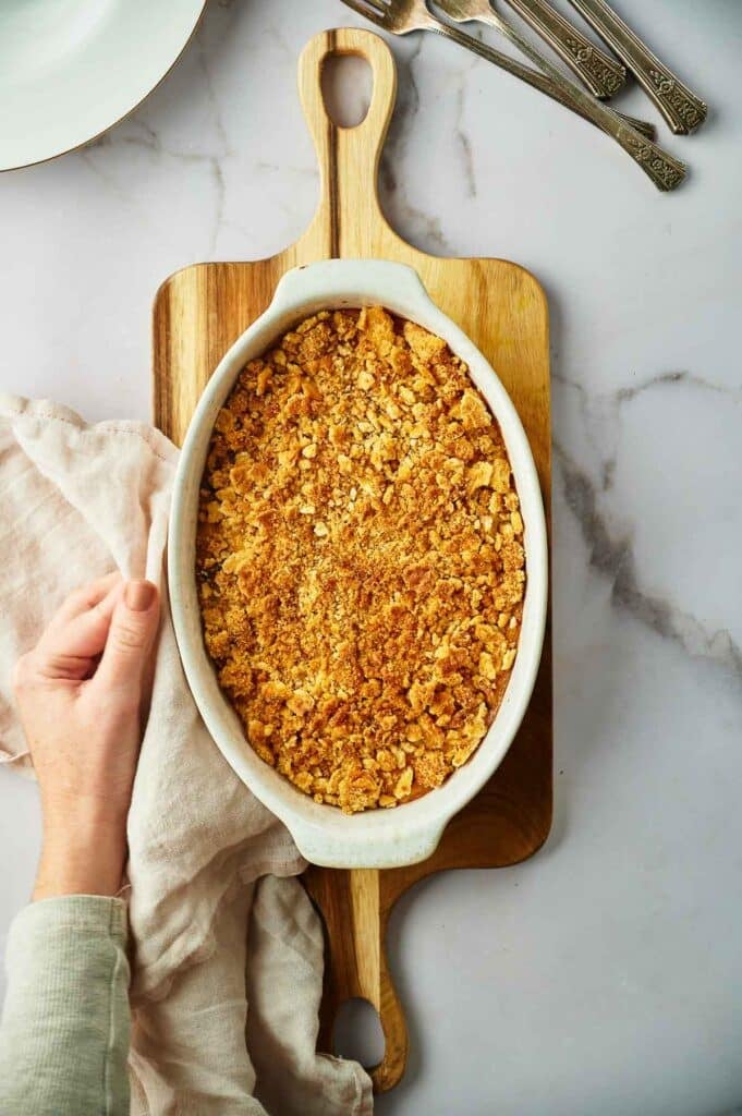 An oval white baking dish of squash casserole with a golden brown crumb topping sits on a wooden cutting board; a hand holds a nearby cloth. Three forks and a plate are in the background.
