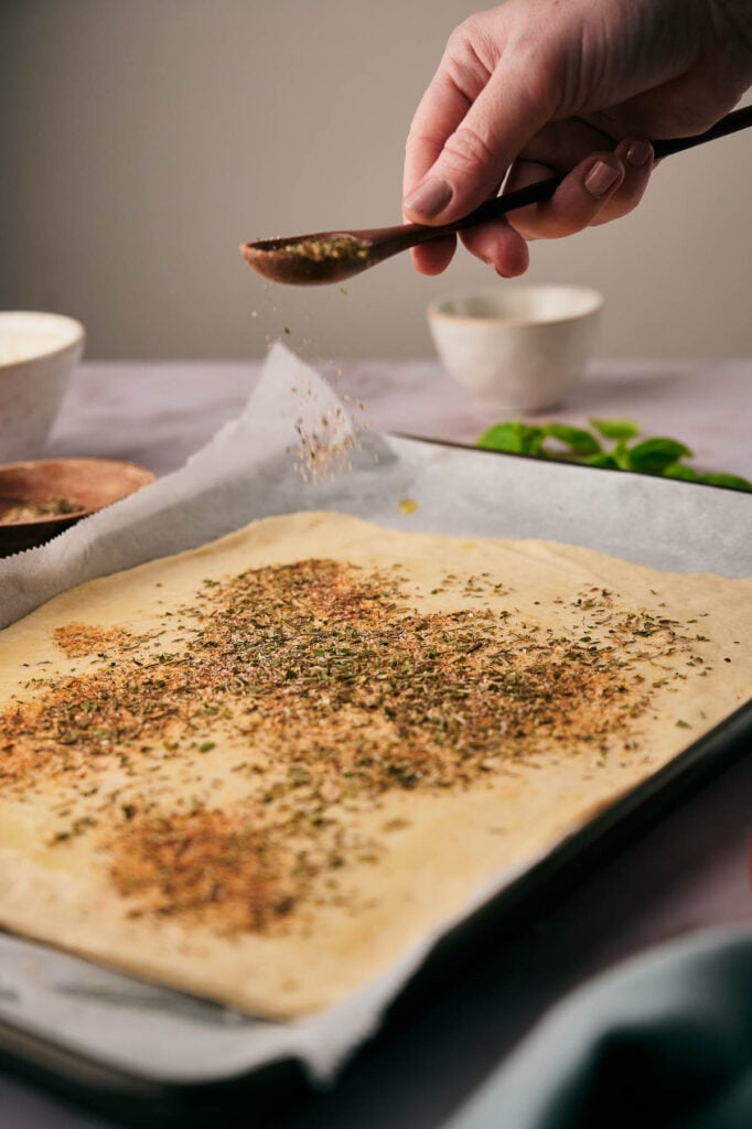 Sprinkling pizza seasoning over pizza dough.
