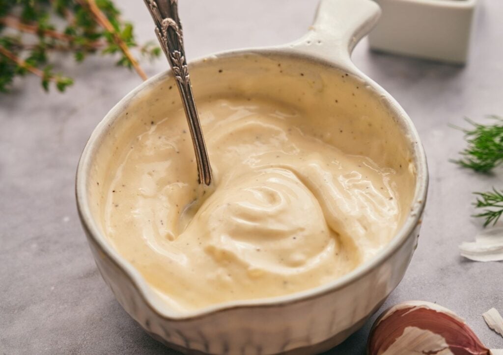 A white ceramic bowl filled with creamy, off-white garlic aioli sits on a gray surface with a spoon inside. Fresh herbs and garlic are nearby.