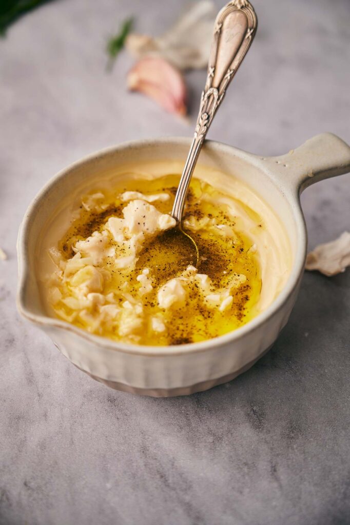 A ceramic bowl filled with mashed potatoes topped with melted butter, cracked black pepper, and a dollop of garlic aioli, with a spoon resting inside.