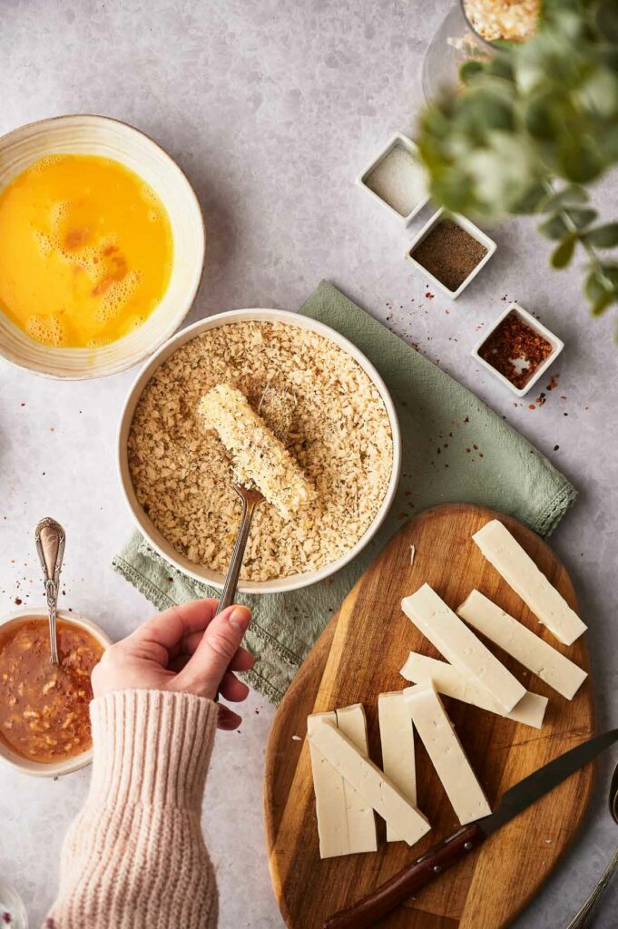 Somone lifting the mozzarella stick out of the breadcrumb mixture.