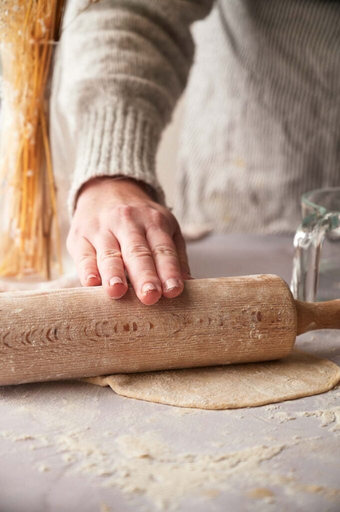 A person rolls out flour tortilla dough with a wooden rolling pin on a floured surface; a bundle of wheat and a glass measuring cup are nearby.