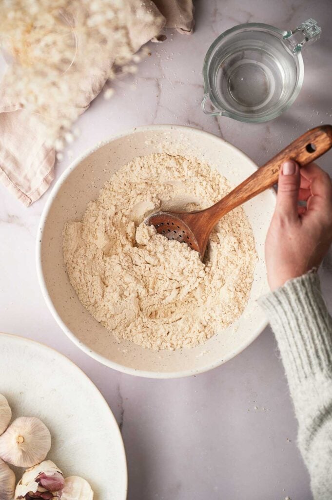A hand stirs flour for flour tortilla dough in a white bowl with a wooden spoon. A glass of water, a beige cloth, and garlic bulbs are nearby on a light-colored surface.