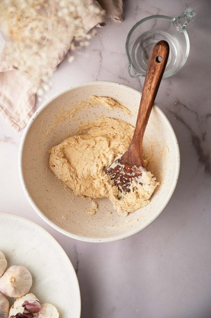 A wooden spoon rests in a bowl of flour tortilla dough on a marble surface, with a glass measuring cup, garlic bulbs, and a cloth nearby.