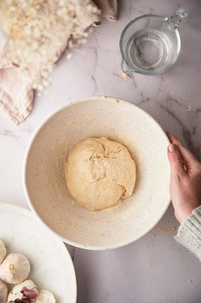 A hand holds a bowl containing a ball of flour tortilla dough on a marble surface, with a glass measuring cup of water and garlic cloves nearby.
