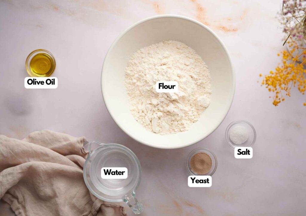 A bowl of flour, with smaller bowls of olive oil, water, yeast, and salt labeled, arranged on a light surface—perfectly prepped for making Ooni pizza dough.