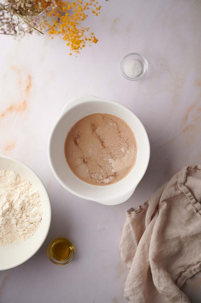A white bowl with yeast and water sits on a countertop next to a plate of flour for Ooni pizza dough, a small glass of oil, salt in a small bowl, a beige cloth, and dried flowers.