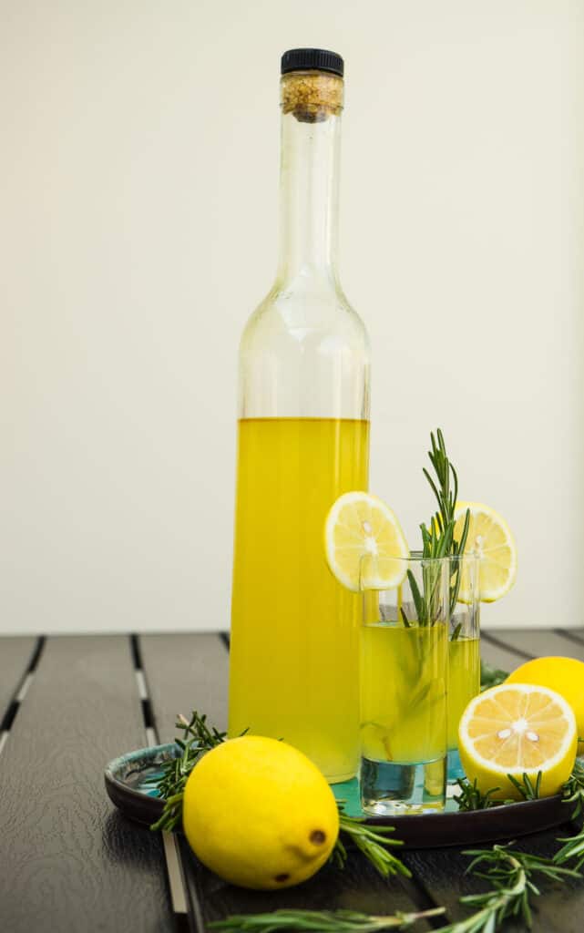 Traditional italian lemon alcohol drink limoncello with pieces of lemon and rosemary herb on dark wooden table