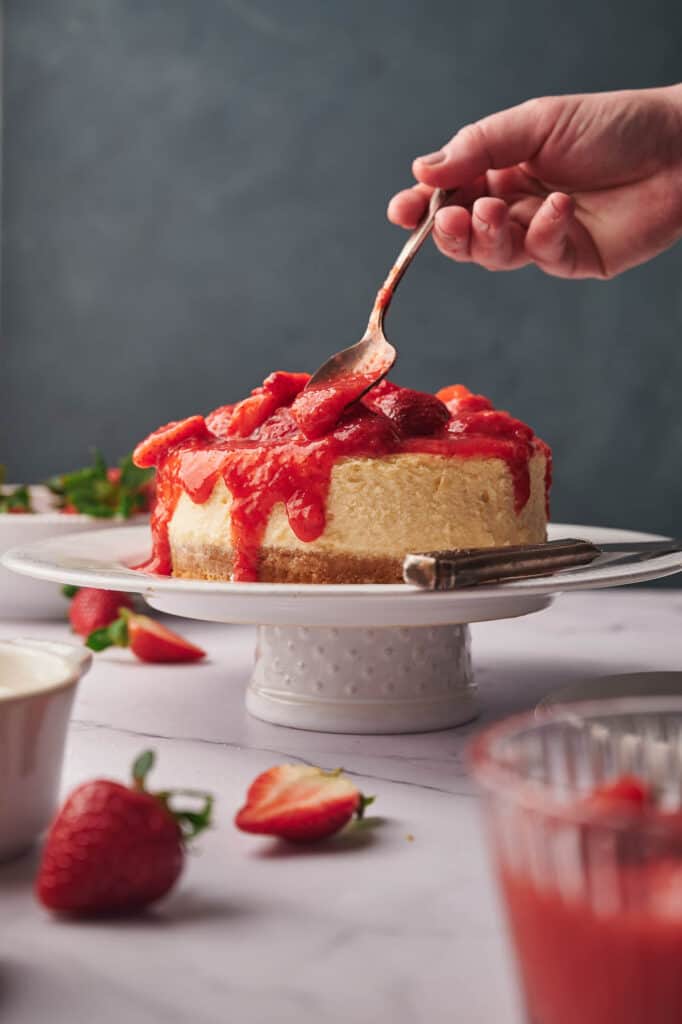 Someone spooning strawberry topping over a baked cheesecake.