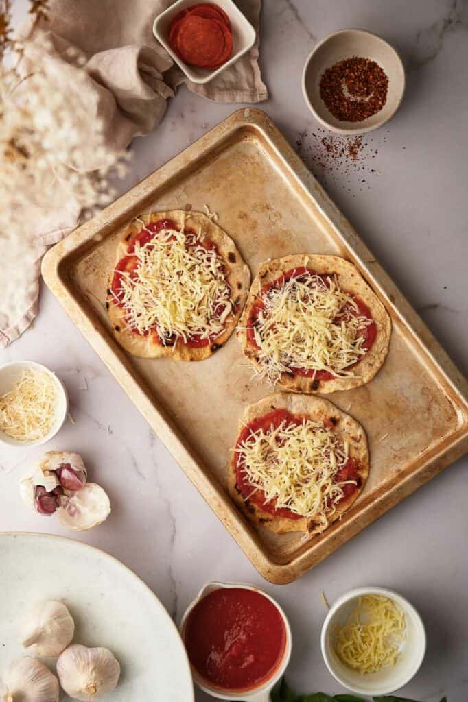 Tortillas on a baking sheet with pizza sauce and shredded mozzarella cheese.