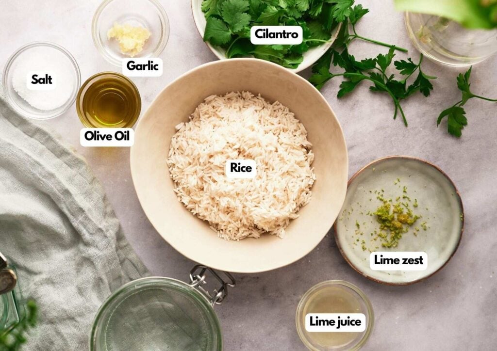 A bowl of uncooked rice is surrounded by labeled ingredients for Cilantro Lime Rice: cilantro, garlic, salt, olive oil, lime zest, and lime juice, all arranged on a light-colored surface.
