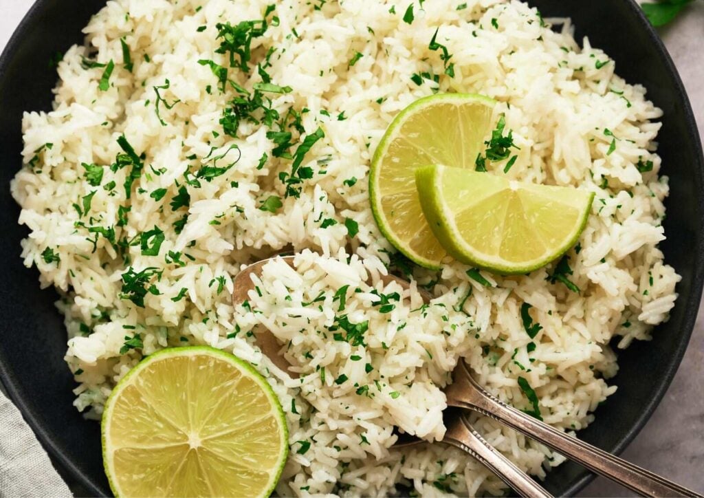 A bowl of Cilantro Lime Rice garnished with chopped herbs and lime wedges, with two spoons resting in the rice.