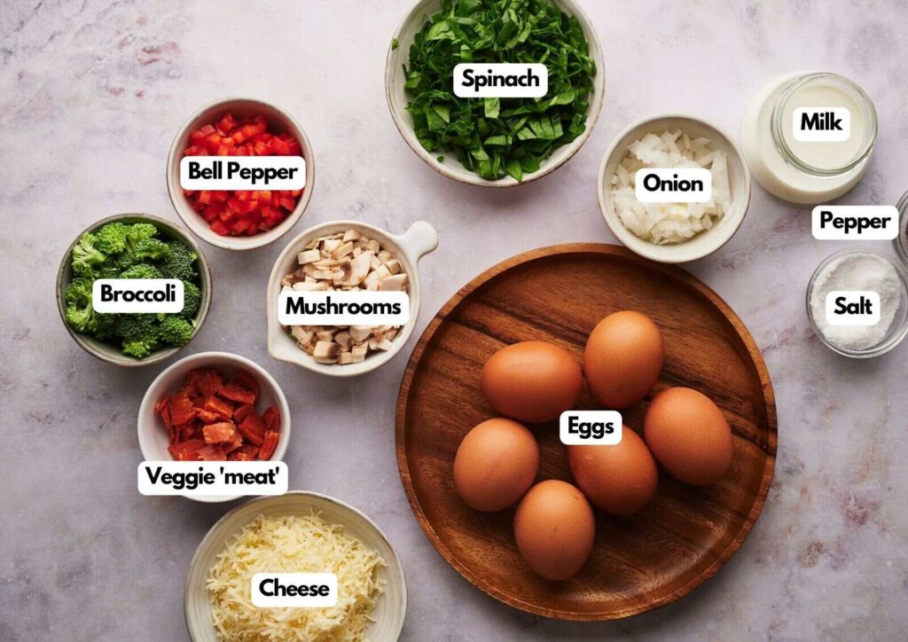 A top view of labeled ingredients for egg bites recipe, including eggs, cheese, spinach, broccoli, bell pepper, onion, mushrooms, veggie “meat,” milk, salt, and pepper on a marble surface.