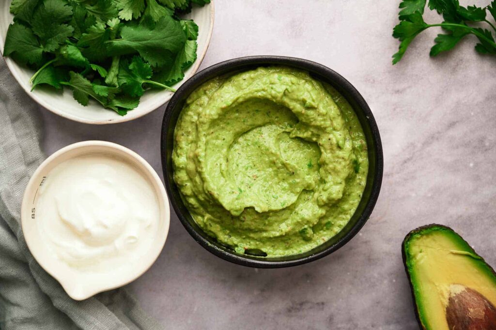 Creamy avocado sauce in a bowl with the ingredients surrounding it.
