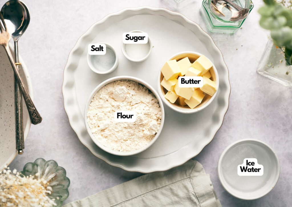 A plate holds bowls labeled flour, butter, salt, and sugar&mdash;with a glass of ice water&mdash;neatly arranged on a light surface with kitchen utensils nearby, ready for your favorite pie crust recipe.