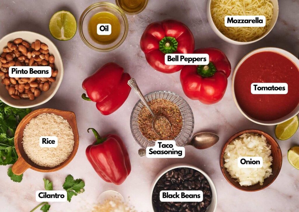 Top view of labeled ingredients in bowls and on a table, including pinto beans, rice, cilantro, oil, bell peppers ready for vegetarian stuffed peppers, taco seasoning, mozzarella, tomatoes, onion, and black beans.