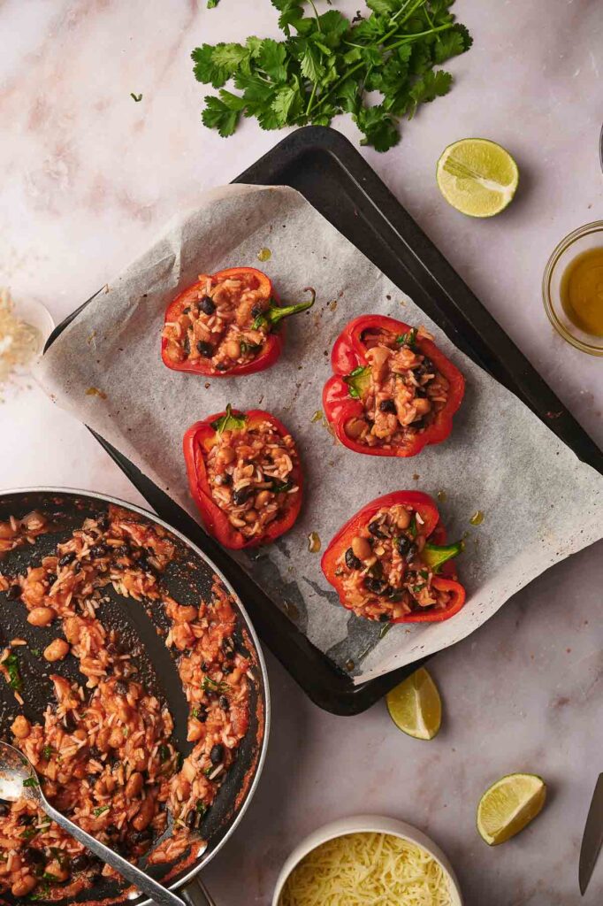 Baking tray with four vegetarian stuffed peppers on parchment paper, a skillet with filling, shredded cheese, lime wedges, and cilantro on a marble surface.