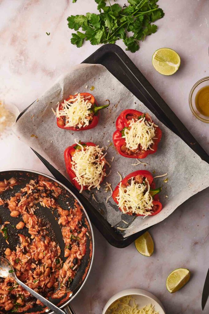 Baking tray with halved bell peppers stuffed with a flavorful vegetarian bean mixture and topped with shredded cheese, beside a pan of filling, lime wedges, cilantro, and olive oil on a marble surface—perfect for serving delicious vegetarian stuffed peppers.