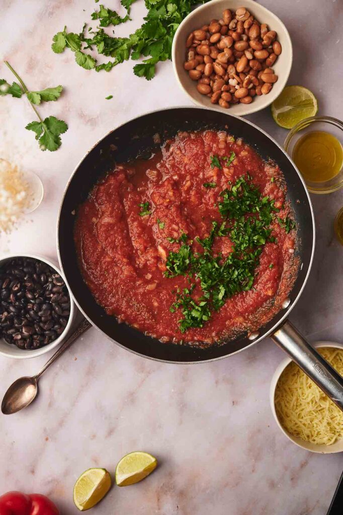 A frying pan with tomato sauce and chopped herbs is surrounded by bowls of beans, cheese, rice, lime wedges, cilantro, and a small glass of oil on a marble surface—the perfect setup for preparing delicious vegetarian stuffed peppers.